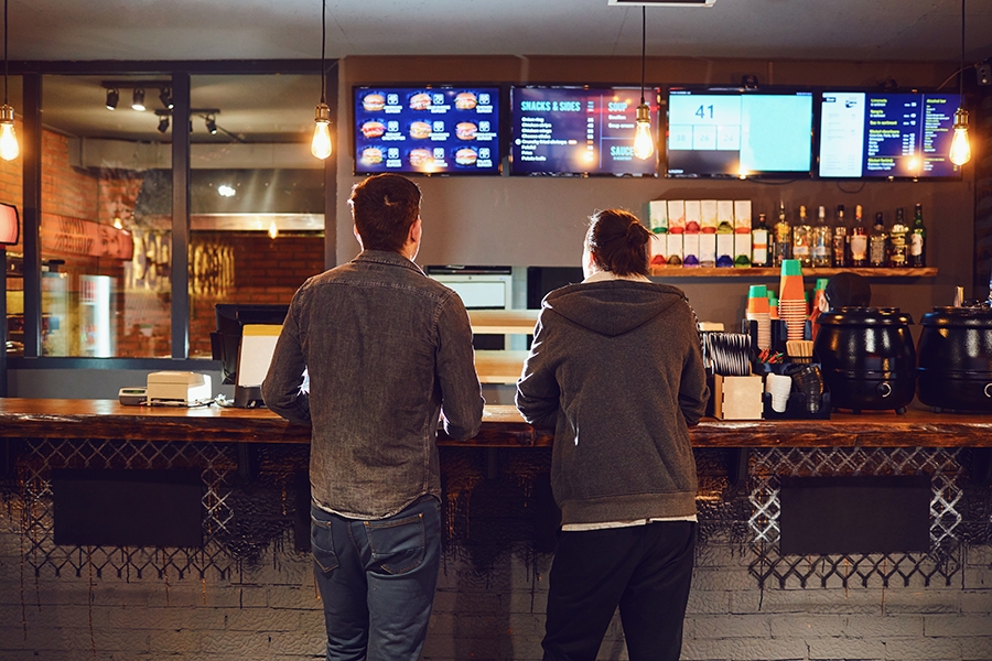 Two people stood at bar infront of TV screens displaying menus and collection points
